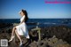 A woman in a white dress sitting on a bench by the ocean.